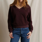 A woman modeling a dark burgundy v-neck knit sweater paired with blue jeans, standing against a neutral draped fabric backdrop.
