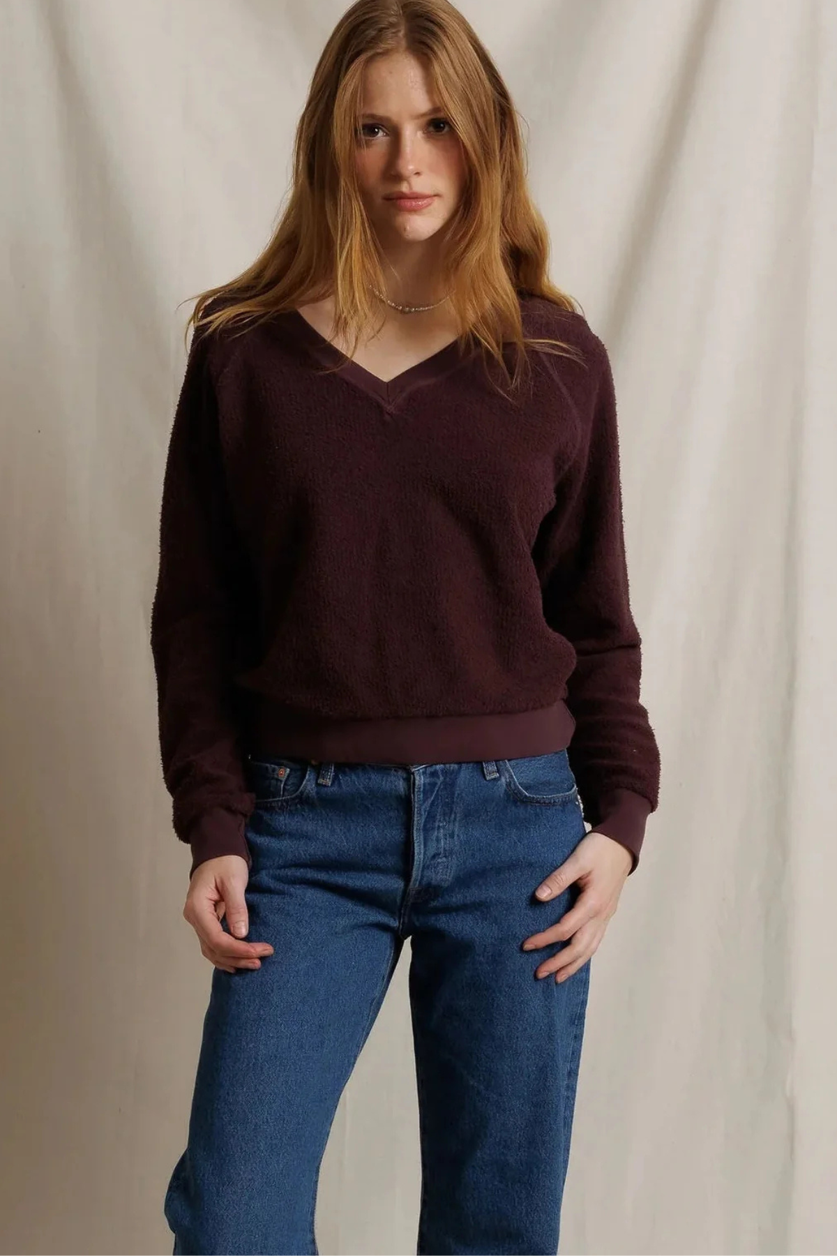 A woman modeling a dark burgundy v-neck knit sweater paired with blue jeans, standing against a neutral draped fabric backdrop.