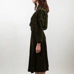 A woman stands in profile wearing a dark olive green velvet midi dress with a wrap-style bodice and long sleeves, paired with white pointed-toe heels, against a plain light gray studio background.