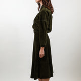 A woman stands in profile wearing a dark olive green velvet midi dress with a wrap-style bodice and long sleeves, paired with white pointed-toe heels, against a plain light gray studio background.