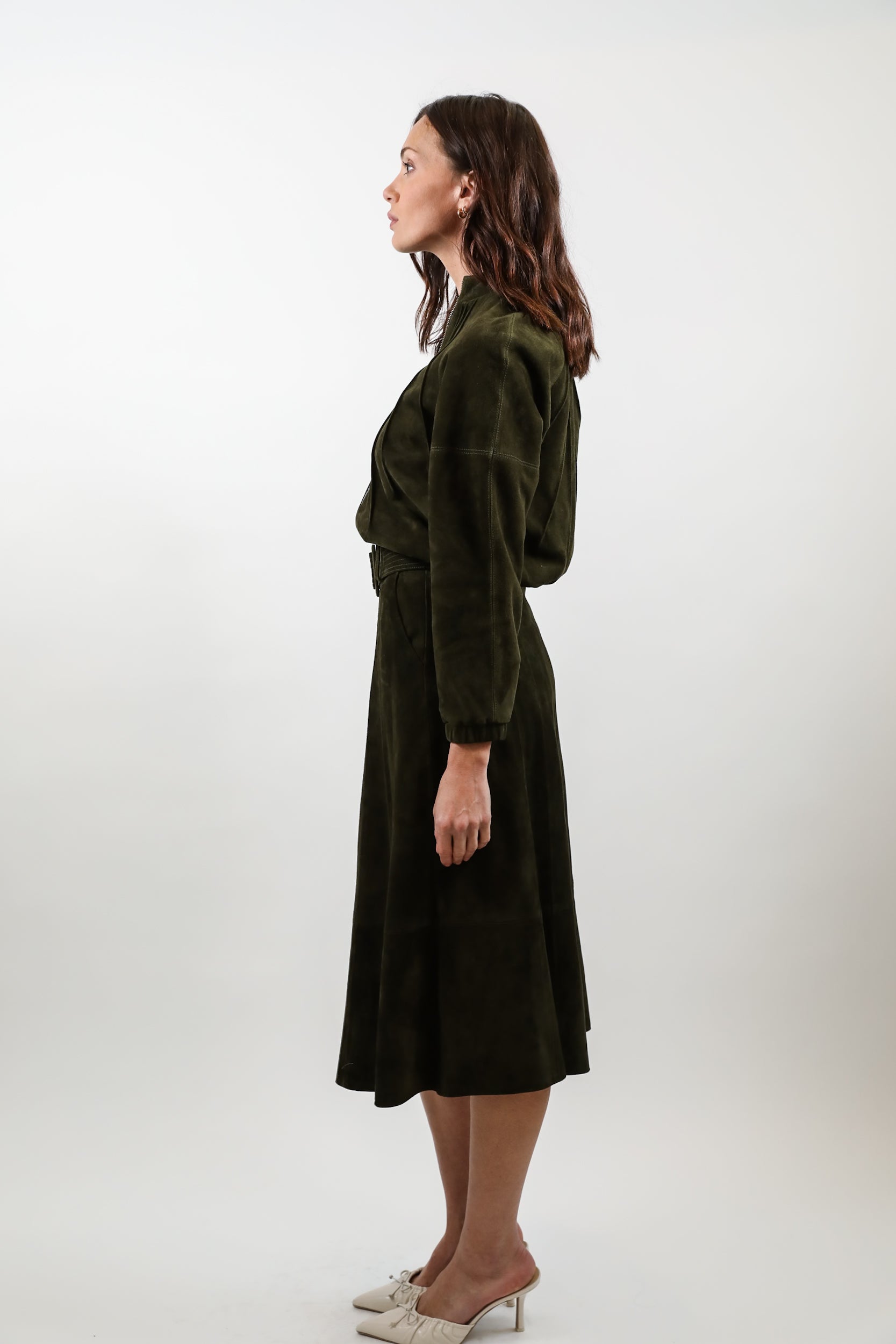 A woman stands in profile wearing a dark olive green velvet midi dress with a wrap-style bodice and long sleeves, paired with white pointed-toe heels, against a plain light gray studio background.