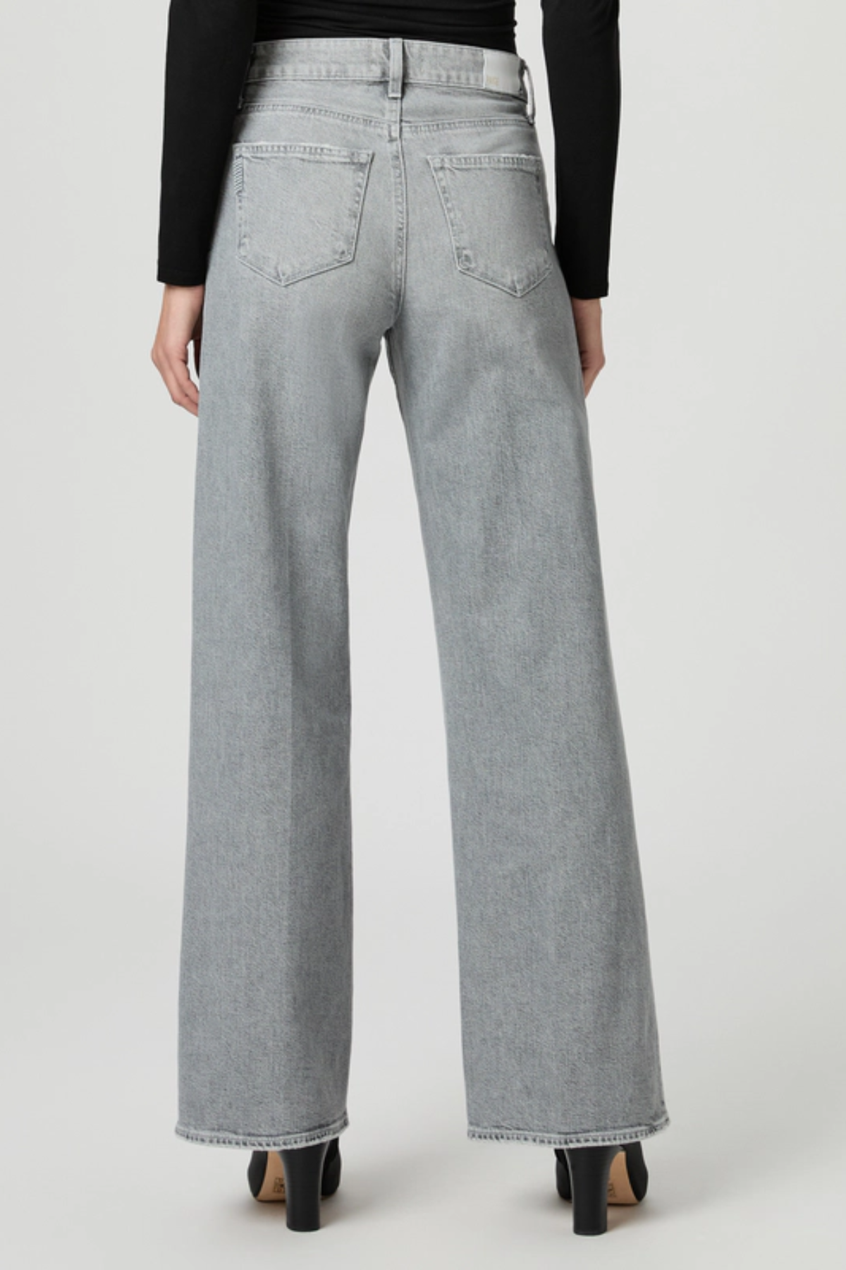 A person shown from the back wearing light gray wide-leg jeans, a black long-sleeve top, and black heels against a plain light gray studio background.