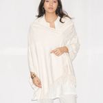 A woman modeling a cream-colored shawl with fringe detailing, worn over a white top and white pants against a plain white studio background.