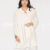 A woman modeling a cream-colored shawl with fringe detailing, worn over a white top and white pants against a plain white studio background.