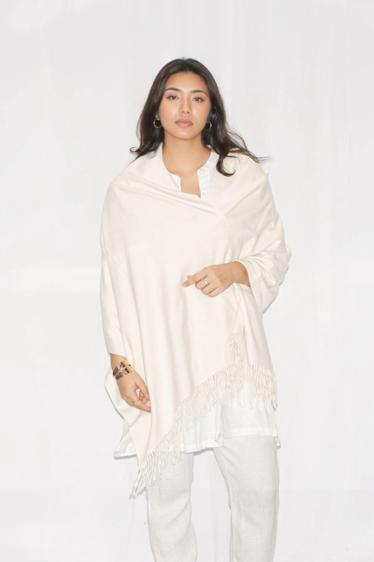 A woman modeling a cream-colored shawl with fringe detailing, worn over a white top and white pants against a plain white studio background.