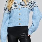 A woman wearing a light blue fuzzy knit cardigan with black and white geometric patterns across the chest and shoulders, accented by gold-tone buttons down the front, paired with black faux-leather pants.
