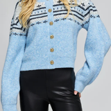 A woman wearing a light blue fuzzy knit cardigan with black and white geometric patterns across the chest and shoulders, accented by gold-tone buttons down the front, paired with black faux-leather pants.