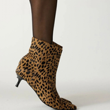 A person wearing a leopard-print ankle boot with a pointed toe and kitten heel, paired with sheer black tights, photographed against a plain light gray studio background.
