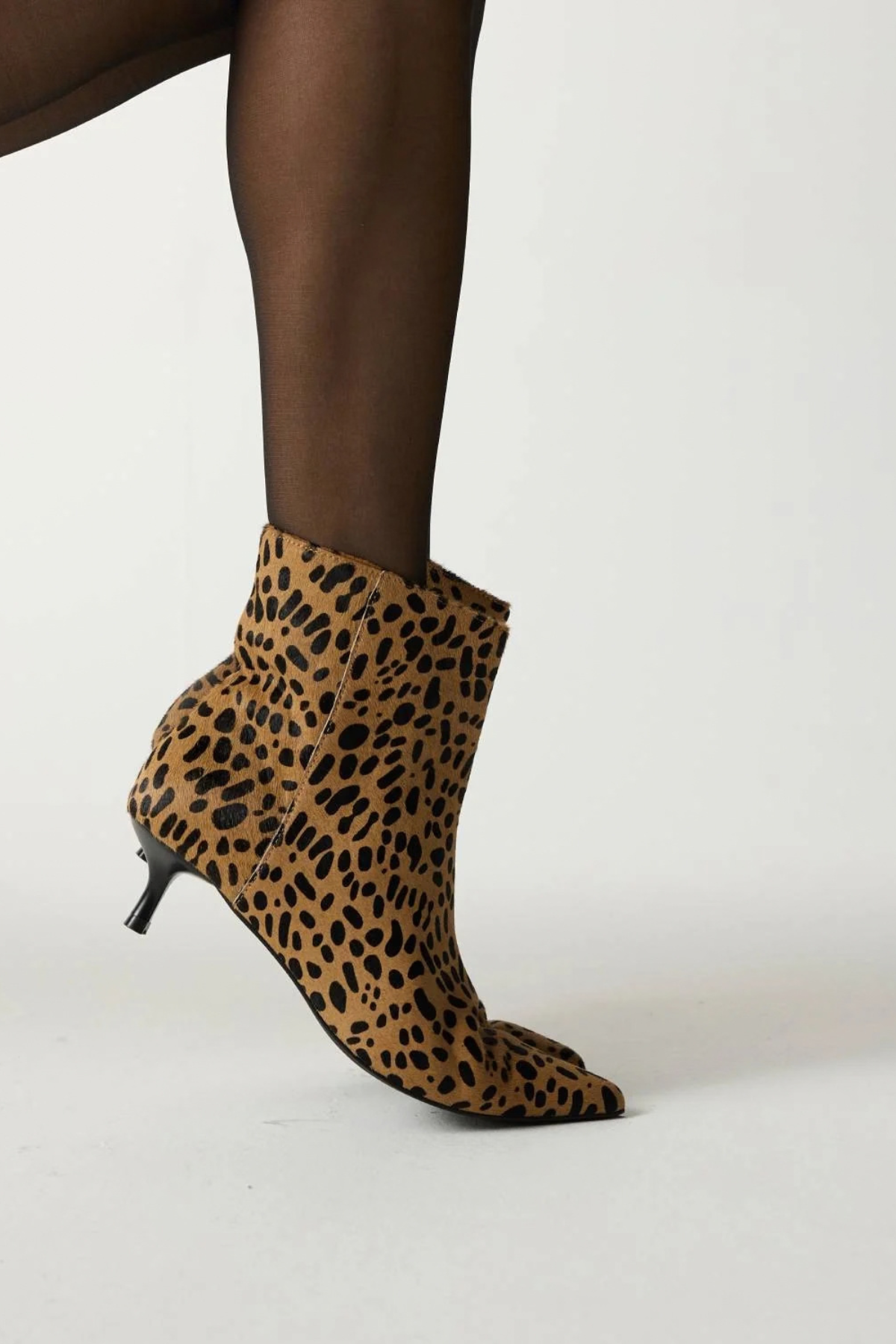 A person wearing a leopard-print ankle boot with a pointed toe and kitten heel, paired with sheer black tights, photographed against a plain light gray studio background.
