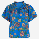 A vibrant blue short-sleeve blouse with a multicolor floral and paisley print, featuring a ruffled high neckline and tie-front detail, shown against a plain light gray background.
