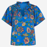 A vibrant blue short-sleeve blouse with a multicolor floral and paisley print, featuring a ruffled high neckline and tie-front detail, shown against a plain light gray background.