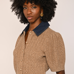 A model with a large black afro wearing a short-sleeve beige cable-knit cropped cardigan with a dark blue denim collar, paired with brown pants, against a plain light beige background.