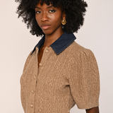 A model with a large black afro wearing a short-sleeve beige cable-knit cropped cardigan with a dark blue denim collar, paired with brown pants, against a plain light beige background.