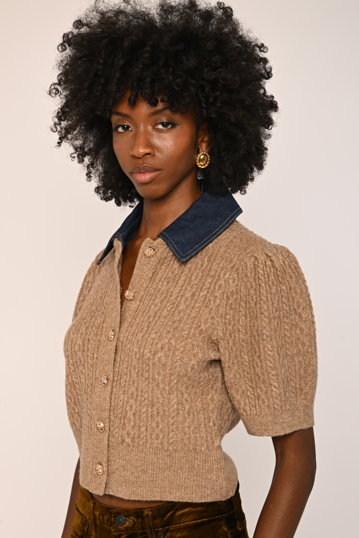 A model with a large black afro wearing a short-sleeve beige cable-knit cropped cardigan with a dark blue denim collar, paired with brown pants, against a plain light beige background.