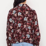 A woman viewed from behind wearing a long-sleeve button-up blouse with a dark burgundy floral print and light blue jeans, set against a plain white background.