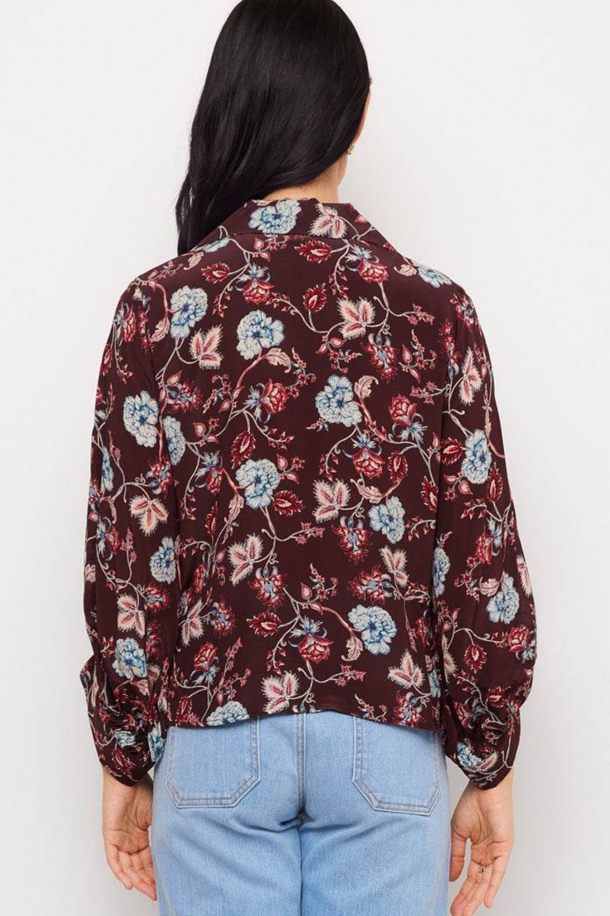 A woman viewed from behind wearing a long-sleeve button-up blouse with a dark burgundy floral print and light blue jeans, set against a plain white background.