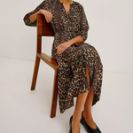 A woman wearing a leopard-print midi dress sits on a wooden chair against a plain white studio background. She is also wearing black pointed flats and dark sunglasses.