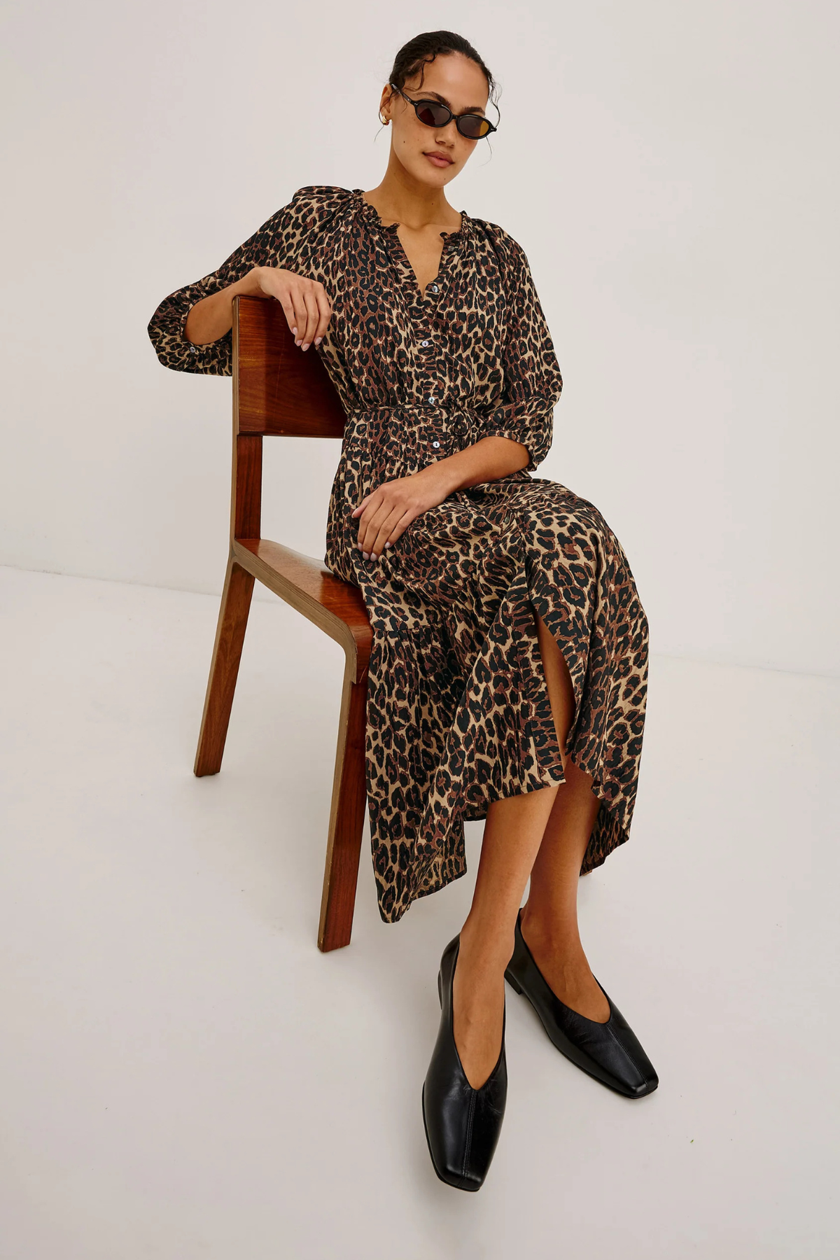 A woman wearing a leopard-print midi dress sits on a wooden chair against a plain white studio background. She is also wearing black pointed flats and dark sunglasses.