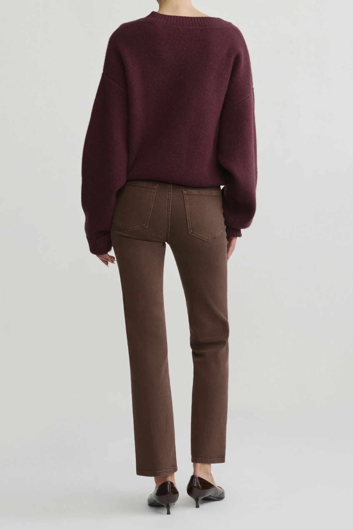 A model shown from the back wearing a dark burgundy knit sweater and brown ankle-length pants, paired with dark brown pointed-toe flats, against a light gray studio background.
