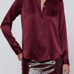 A woman wearing a long-sleeve satin button-up blouse in deep burgundy, paired with shiny dark burgundy crocodile-embossed pants, against a light gray studio background.