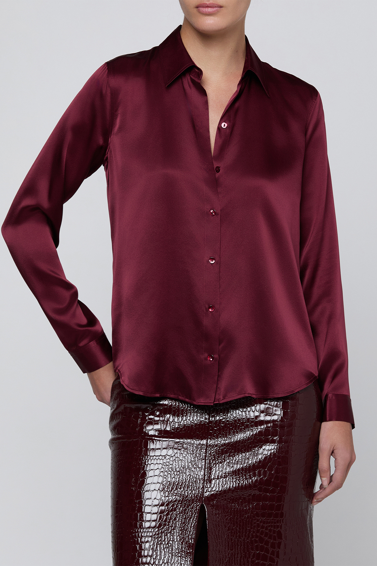 A woman wearing a long-sleeve satin button-up blouse in deep burgundy, paired with shiny dark burgundy crocodile-embossed pants, against a light gray studio background.