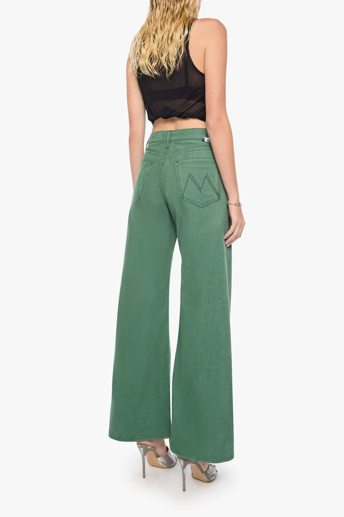 A woman shown from the back wearing green wide-leg pants, a black sleeveless crop top, and metallic high heels against a plain light gray studio background.