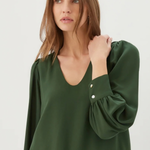 A woman wearing a dark green long-sleeve blouse with puffed sleeves and button details on the cuffs, paired with blue jeans, against a plain light gray background.