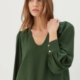 A woman wearing a dark green long-sleeve blouse with puffed sleeves and button details on the cuffs, paired with blue jeans, against a plain light gray background.