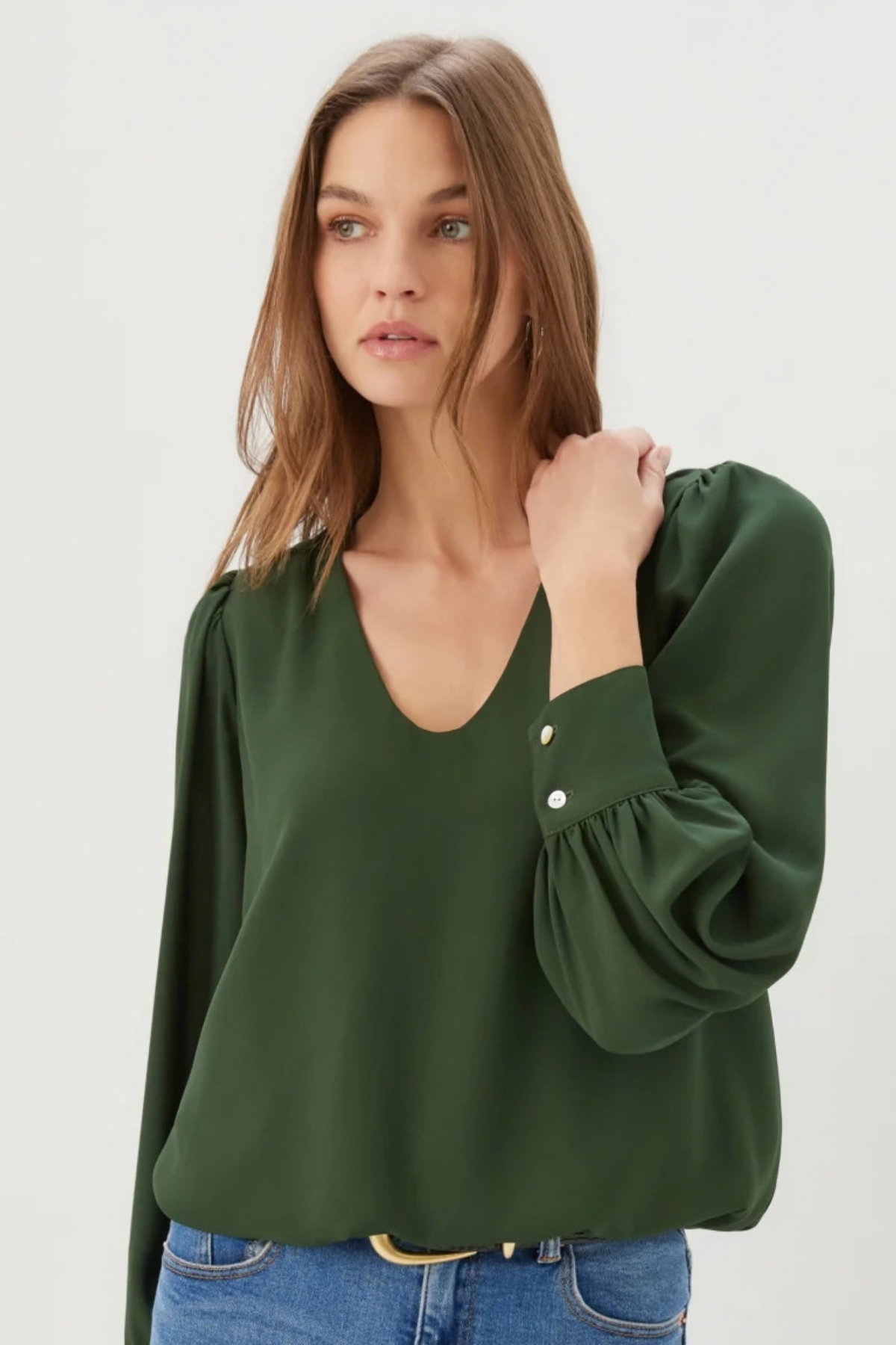A woman wearing a dark green long-sleeve blouse with puffed sleeves and button details on the cuffs, paired with blue jeans, against a plain light gray background.