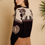 A woman wearing a cropped knit sweater with a cream upper section featuring floral and geometric patterns, and a black lower section, paired with brown pants against a beige background with plant leaves.