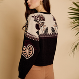 A woman wearing a cropped knit sweater with a cream upper section featuring floral and geometric patterns, and a black lower section, paired with brown pants against a beige background with plant leaves.