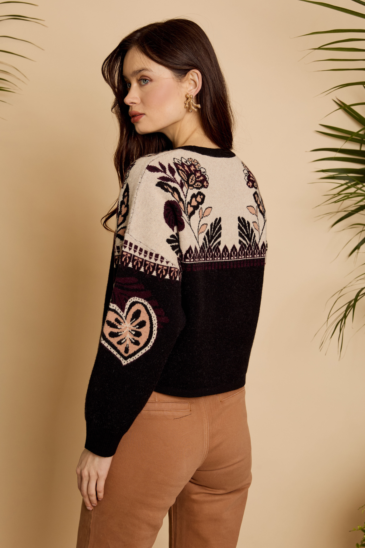 A woman wearing a cropped knit sweater with a cream upper section featuring floral and geometric patterns, and a black lower section, paired with brown pants against a beige background with plant leaves.