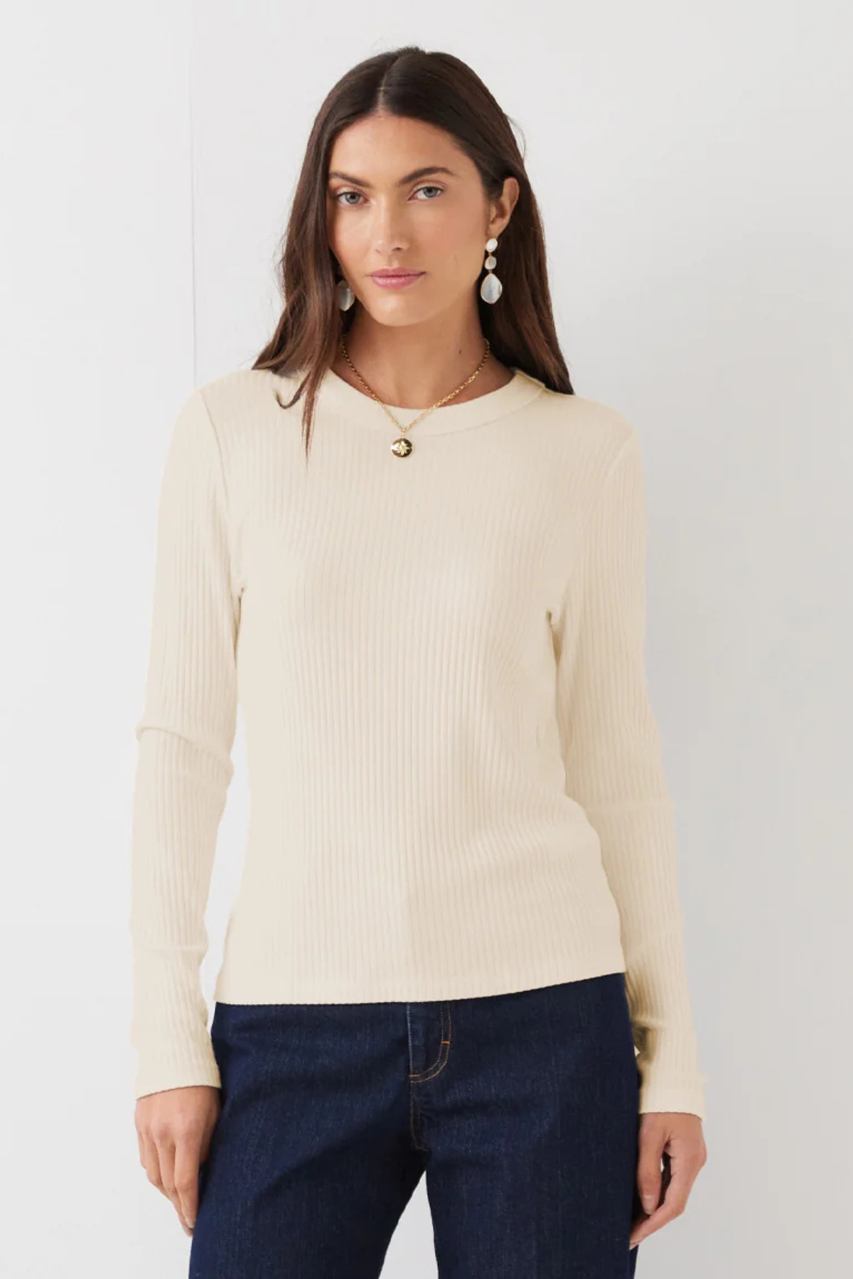 A woman modeling a cream ribbed long-sleeve knit top with dark blue jeans, standing against a plain white background.
