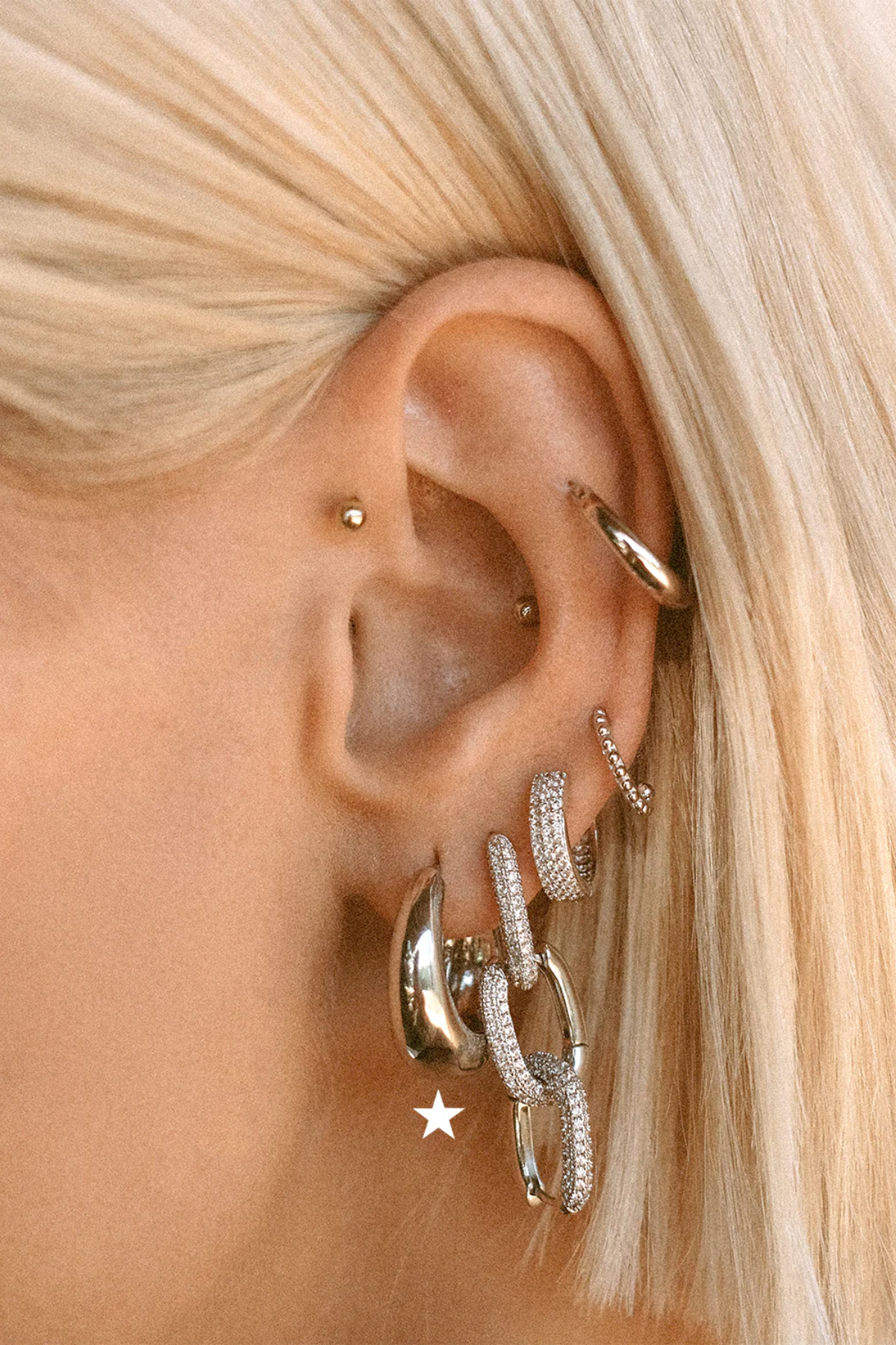 A close-up of a person's ear adorned with multiple silver-toned earrings, including a large chain-link style hoop, a textured ring, a small stud, and a huggie hoop, against blonde hair.