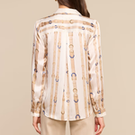 A woman shown from the back wearing a cream satin shirt with vertical gold and blue chain-link patterns, paired with beige trousers, against a solid beige background.