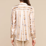 A woman shown from the back wearing a cream satin shirt with vertical gold and blue chain-link patterns, paired with beige trousers, against a solid beige background.