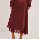 A woman wearing a long-sleeve satin midi skirt in deep burgundy with a lace hem, paired with black strappy heels, shown from the back against a plain light background.