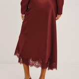 A woman wearing a long-sleeve satin midi skirt in deep burgundy with a lace hem, paired with black strappy heels, shown from the back against a plain light background.
