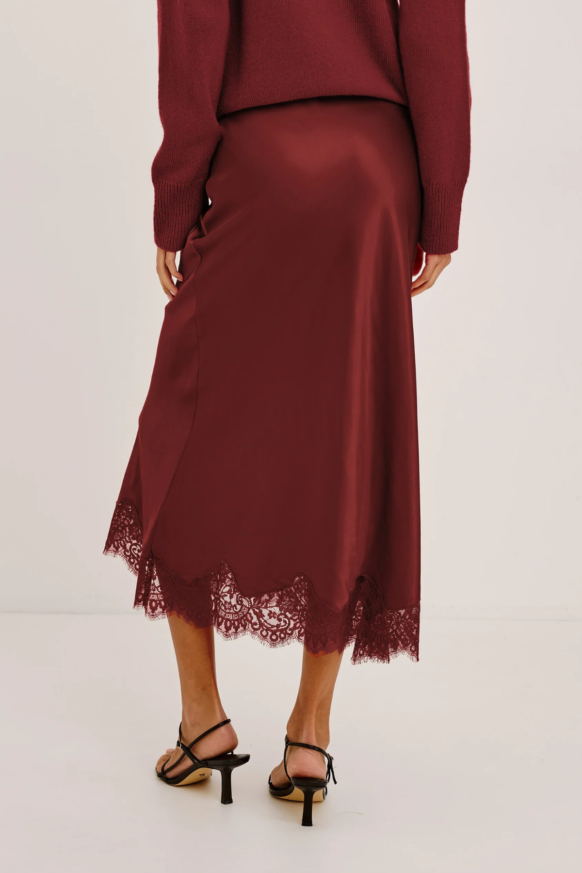 A woman wearing a long-sleeve satin midi skirt in deep burgundy with a lace hem, paired with black strappy heels, shown from the back against a plain light background.