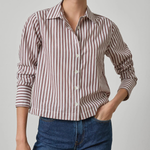 A woman wearing a long-sleeve button-up shirt with vertical brown and white stripes, paired with blue jeans, photographed against a light gray studio background.