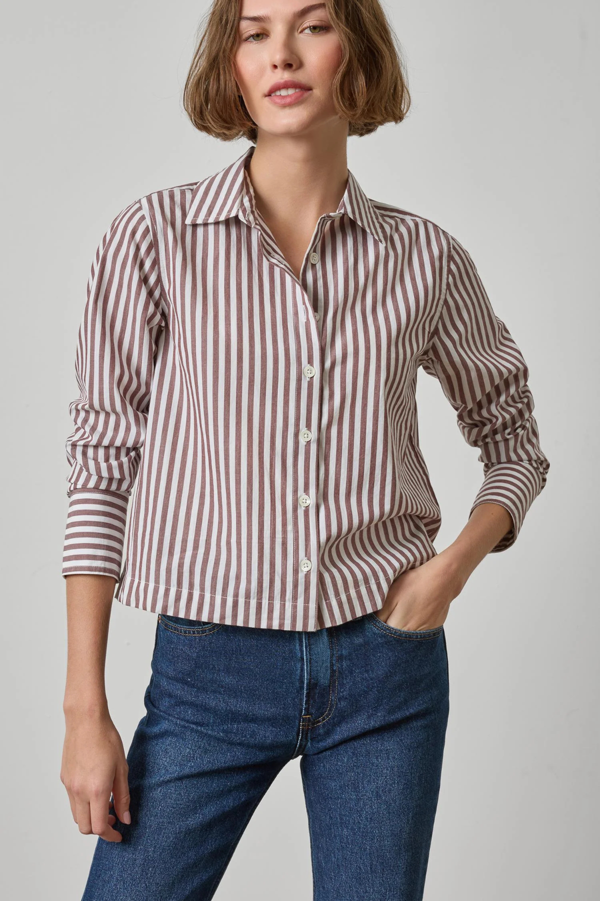A woman wearing a long-sleeve button-up shirt with vertical brown and white stripes, paired with blue jeans, photographed against a light gray studio background.