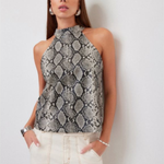 A woman modeling a sleeveless high-neck top with a gray and black snakeskin pattern, paired with cream-colored pants, standing against a plain light gray wall.