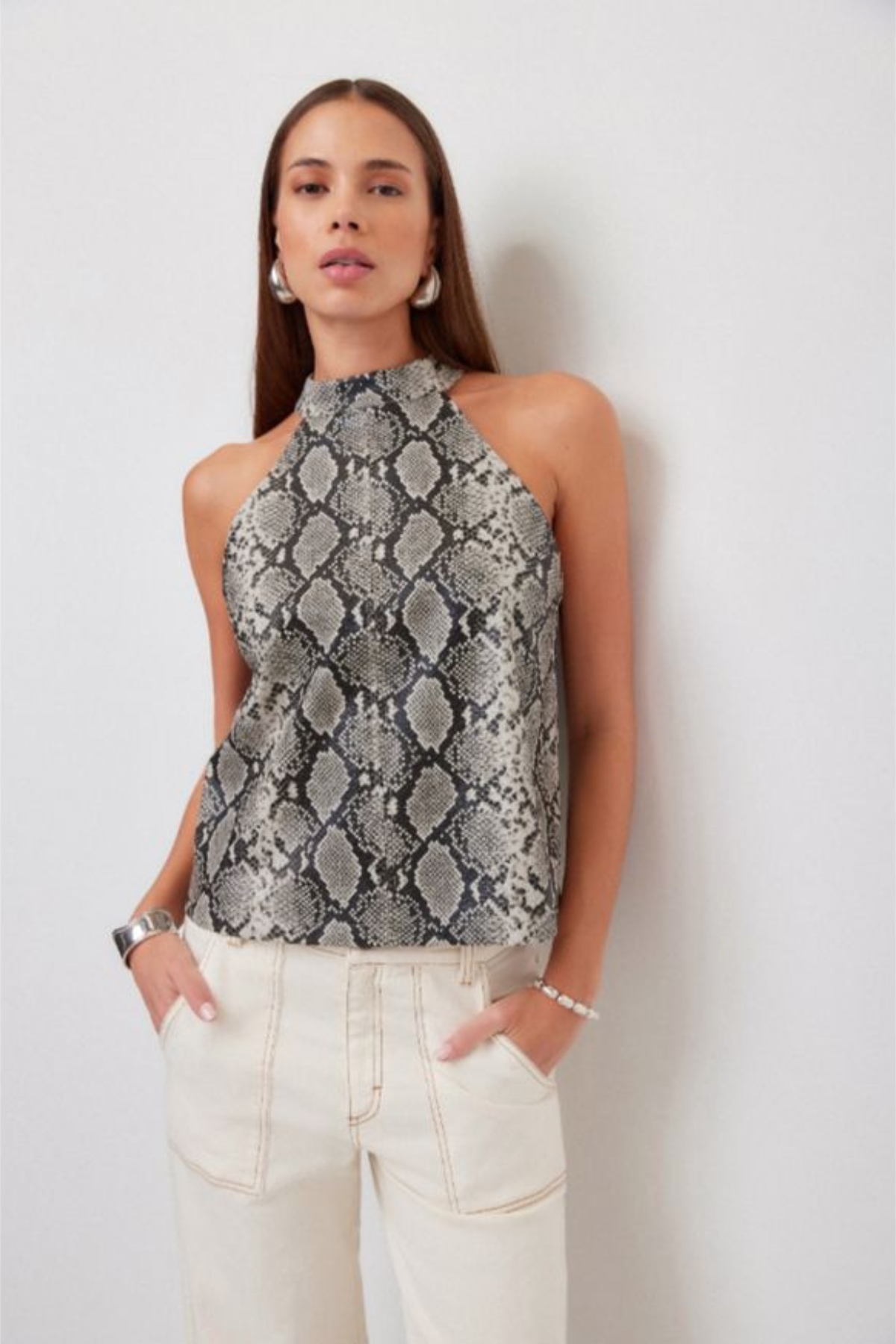 A woman modeling a sleeveless high-neck top with a gray and black snakeskin pattern, paired with cream-colored pants, standing against a plain light gray wall.