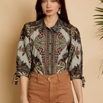 A woman modeling a long-sleeve button-up shirt with a detailed botanical and bird pattern in earthy tones, paired with brown pants, set against a warm beige background with soft plant shadows.