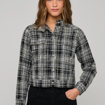 A woman modeling a black and white plaid tweed button-up shirt jacket with black pants, photographed against a solid light gray studio background.