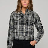 A woman modeling a black and white plaid tweed button-up shirt jacket with black pants, photographed against a solid light gray studio background.
