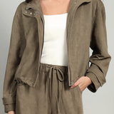 A woman wearing an olive brown suede jacket over a white tank top and matching olive brown drawstring pants, photographed against a light gray studio background.
