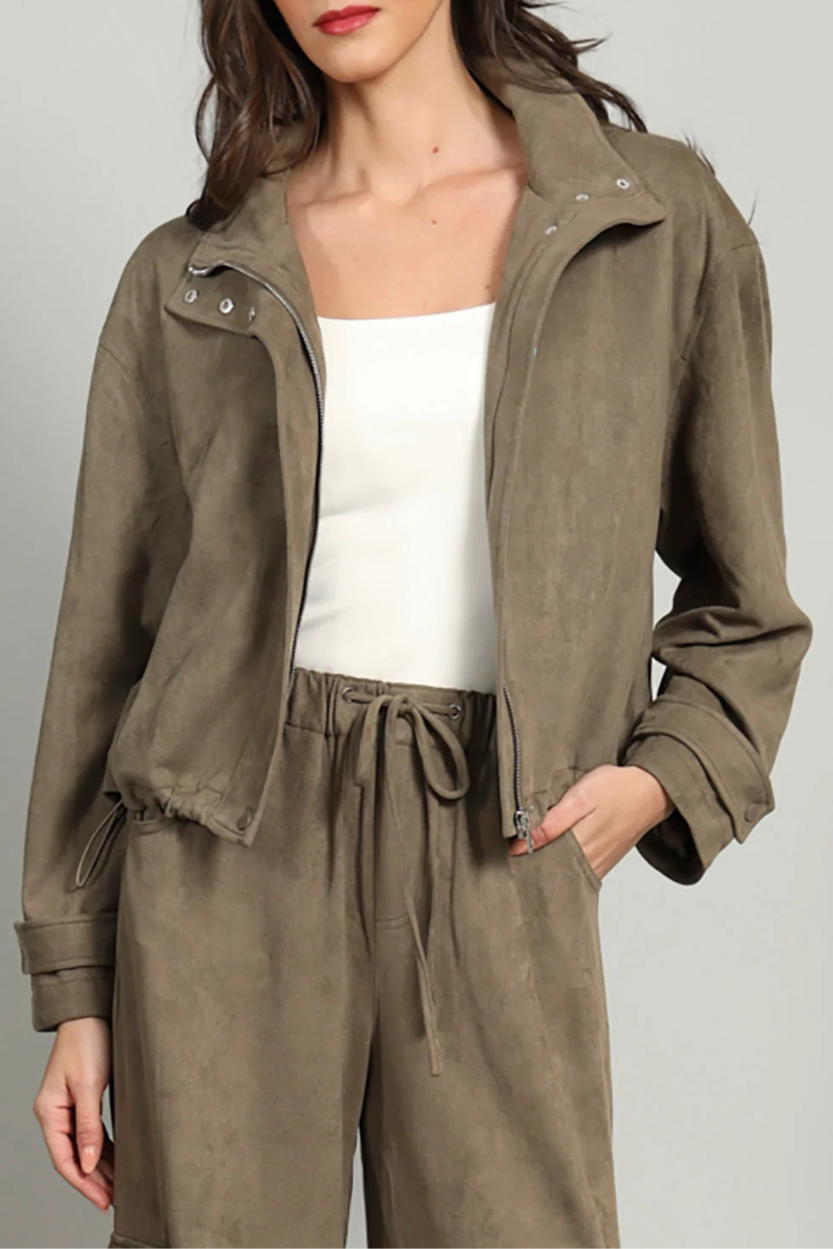 A woman wearing an olive brown suede jacket over a white tank top and matching olive brown drawstring pants, photographed against a light gray studio background.