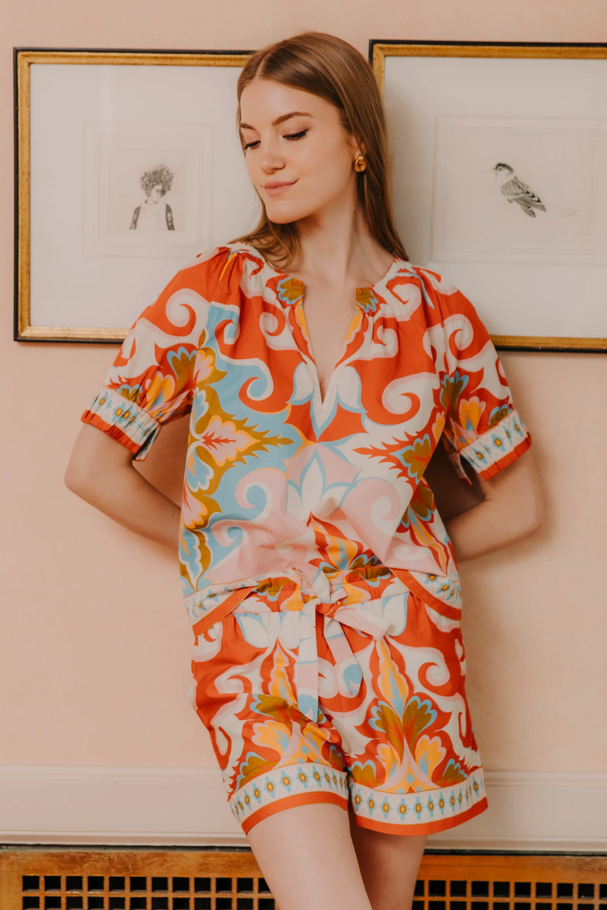 Woman wearing a colorful patterned outfit standing against a light pink wall with framed pictures.