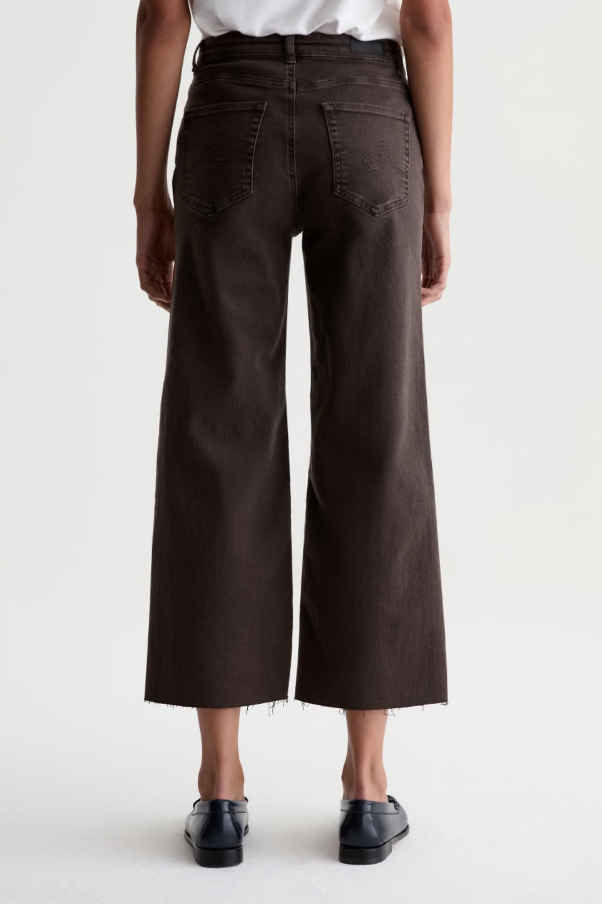 A person shown from the back wearing dark brown wide-leg cropped jeans with a white top and black shoes, photographed against a clean light gray studio background.